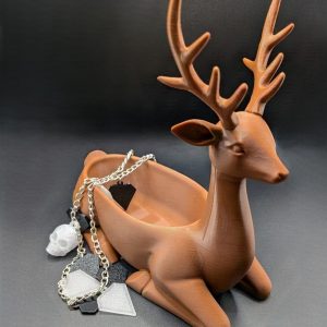 Brown 3D-printed deer storage figure with large antlers, holding silver and black necklaces and pendants, against a dark background.
