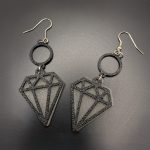 Black geometric diamond dangle earrings suspended from circular accents with silver fish hook fastenings on a dark background.